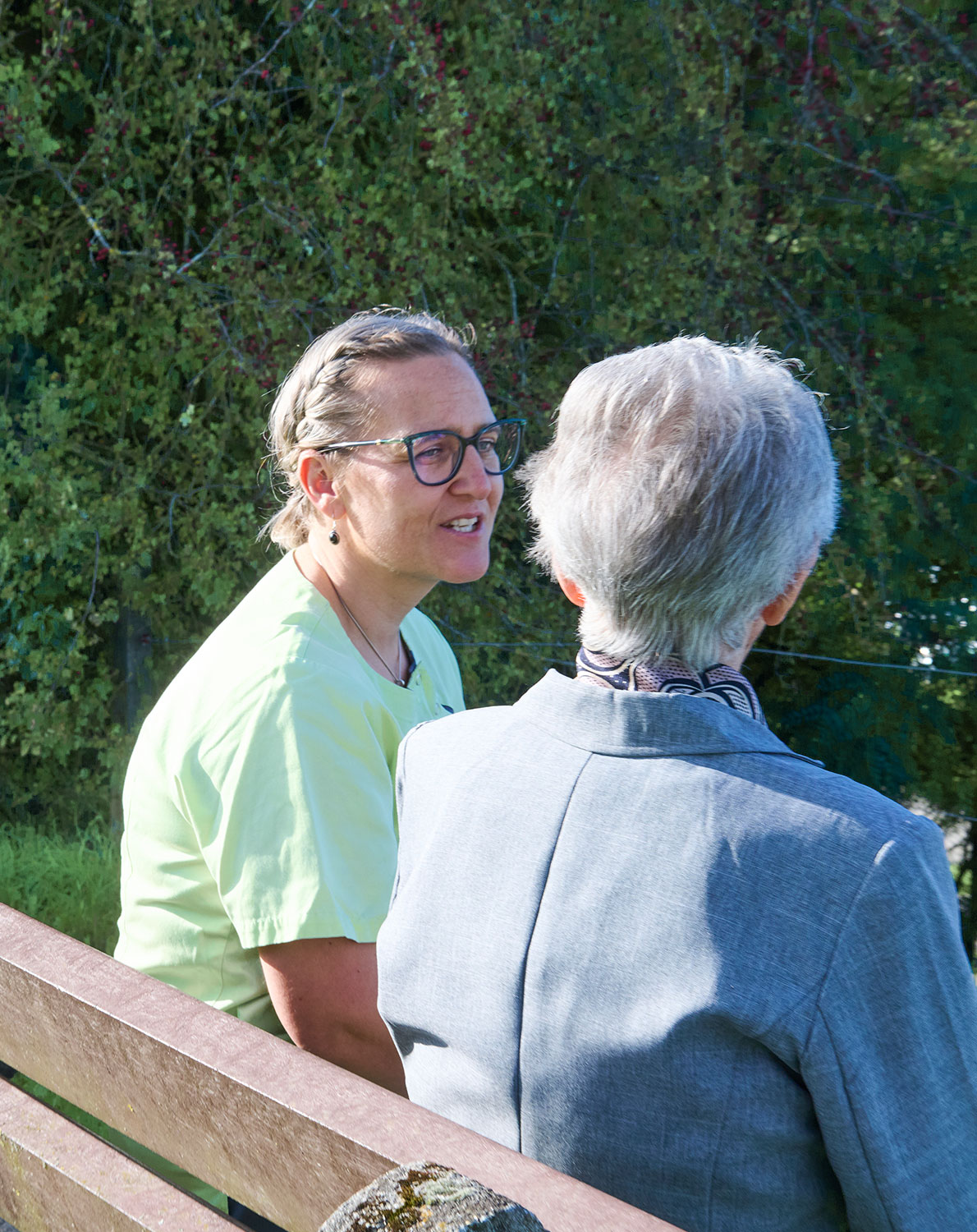 Kontaktaufnahme Eine Pflegerin und eine Bewohnerin sitzen auf einer Gartenbank im Freien und unterhalten sich