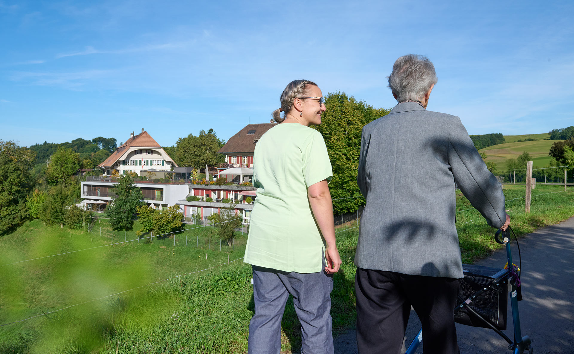Pflege und Betreuung am Landhaus Neuenegg Eine Bewohnerin mit Rollator spaziert mit einer Pflegerin, im Hintergrund die ländliche Umgebung des Landhaus Neuenegg